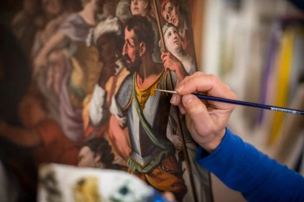 Una mano che dipinge su una tela con un pennello, rappresentando personaggi storici o religiosi in stile realistico.