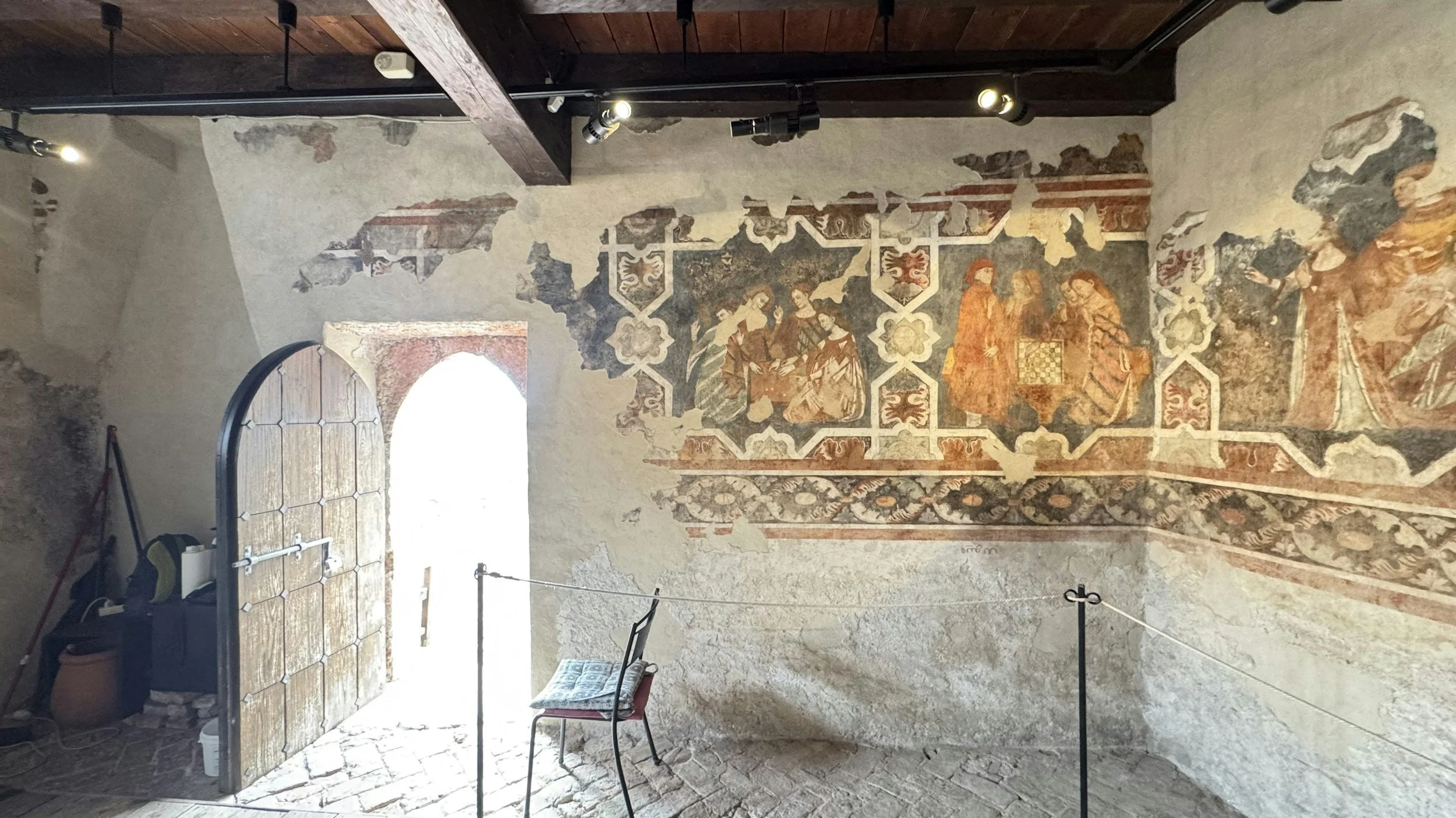 Mura antiche decorate con affreschi religiosi e geometrici, con un'apertura in pietra e legno, e alcuni arredi come sedia, borsa e scalpelli, all'interno di una stanza.