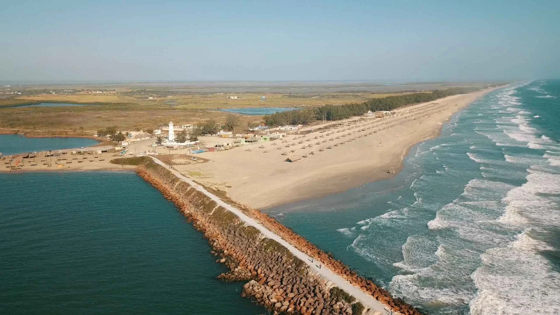 An aerial view of a coastal beach with a breakwater, a lighthouse, and a parking area with beach chairs. The ocean waves are hitting the shore on the right side, and there are small lakes and open land in the background.