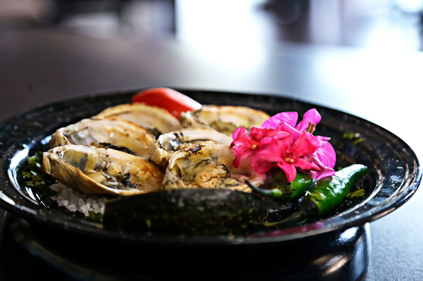 A black plate with grilled oysters topped with herbs, garnished with pink bougainvillea flowers, green chili, and a red chili, set on salt under dim lighting.