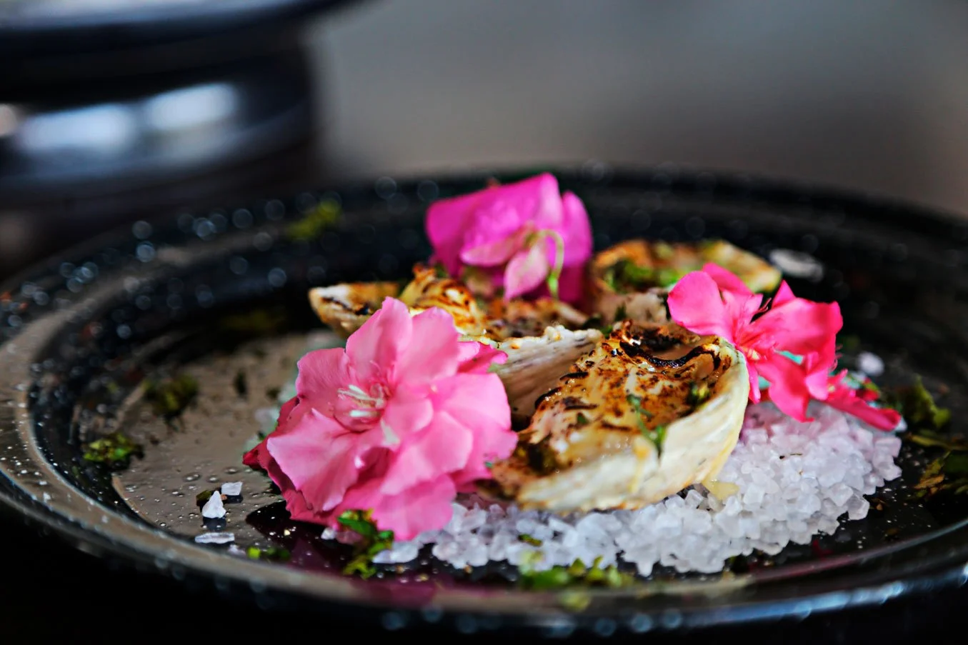 Cooked oysters garnished with pink flowers, served on a bed of salt with herbs.