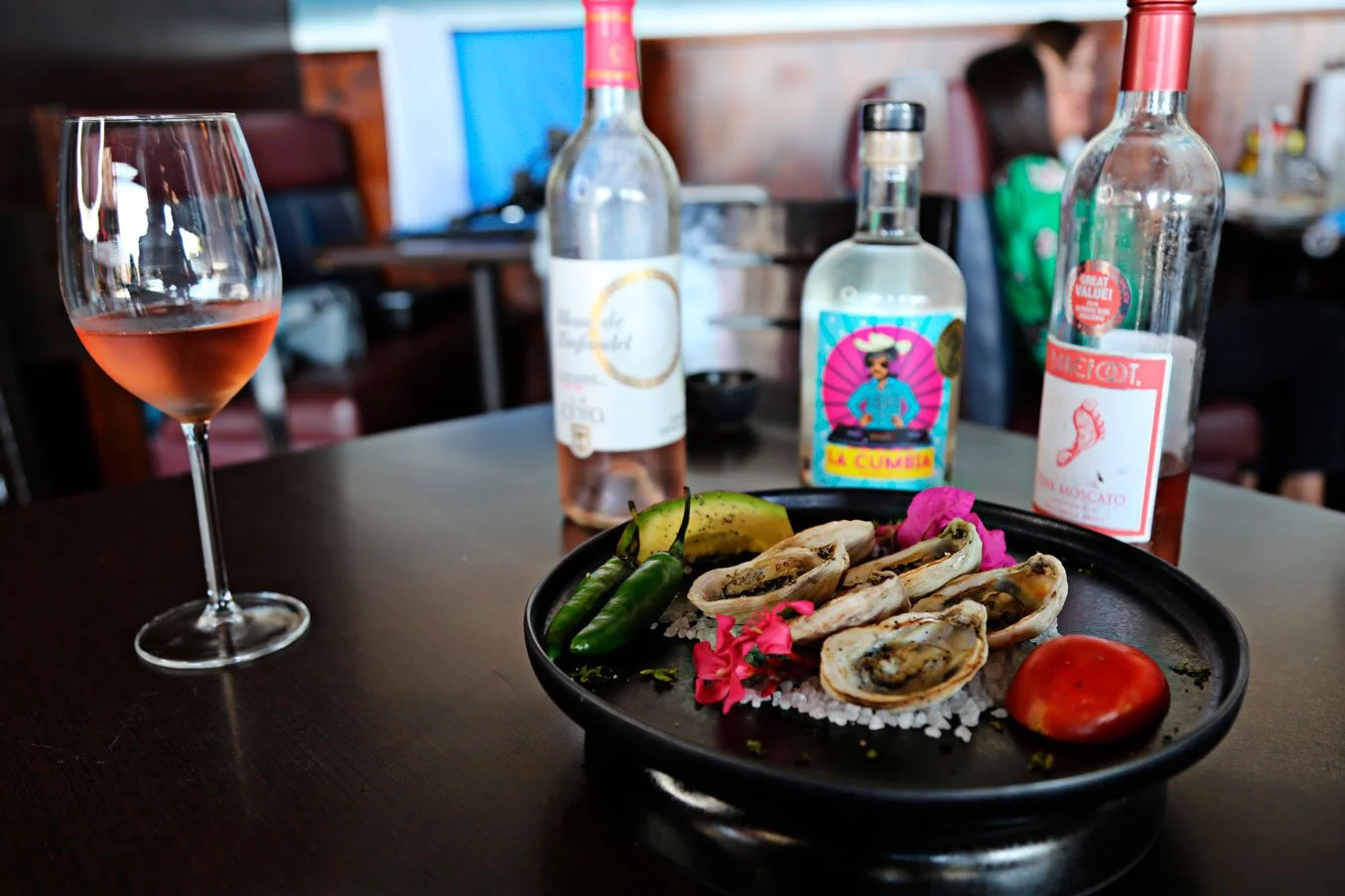 A plate of oysters with lemon, chili peppers, a tomato, and pink flowers on a black plate at a restaurant table. A glass of rosé wine, a partially consumed bottle of rosé wine, a bottle of tequila, and a bottle of mezcal are also on the table.