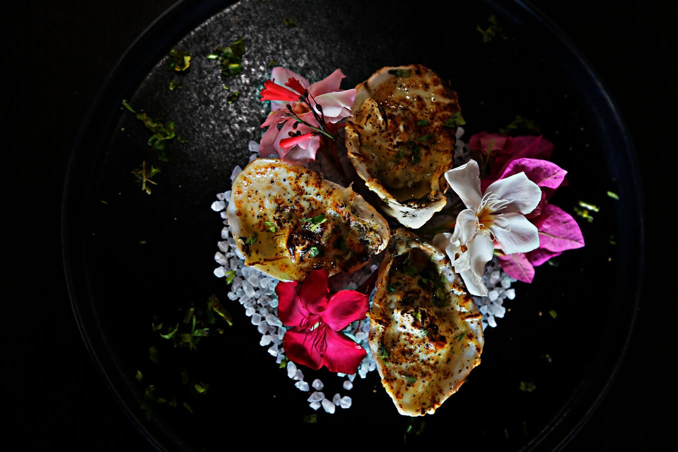 Three oysters topped with sauce and herbs, garnished with edible flowers and sea salt on a black plate.