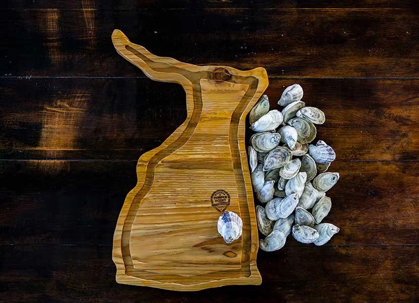 A wooden cutting board in the shape of the state of Georgia, with a pile of raw oysters next to it on a dark wooden surface.
