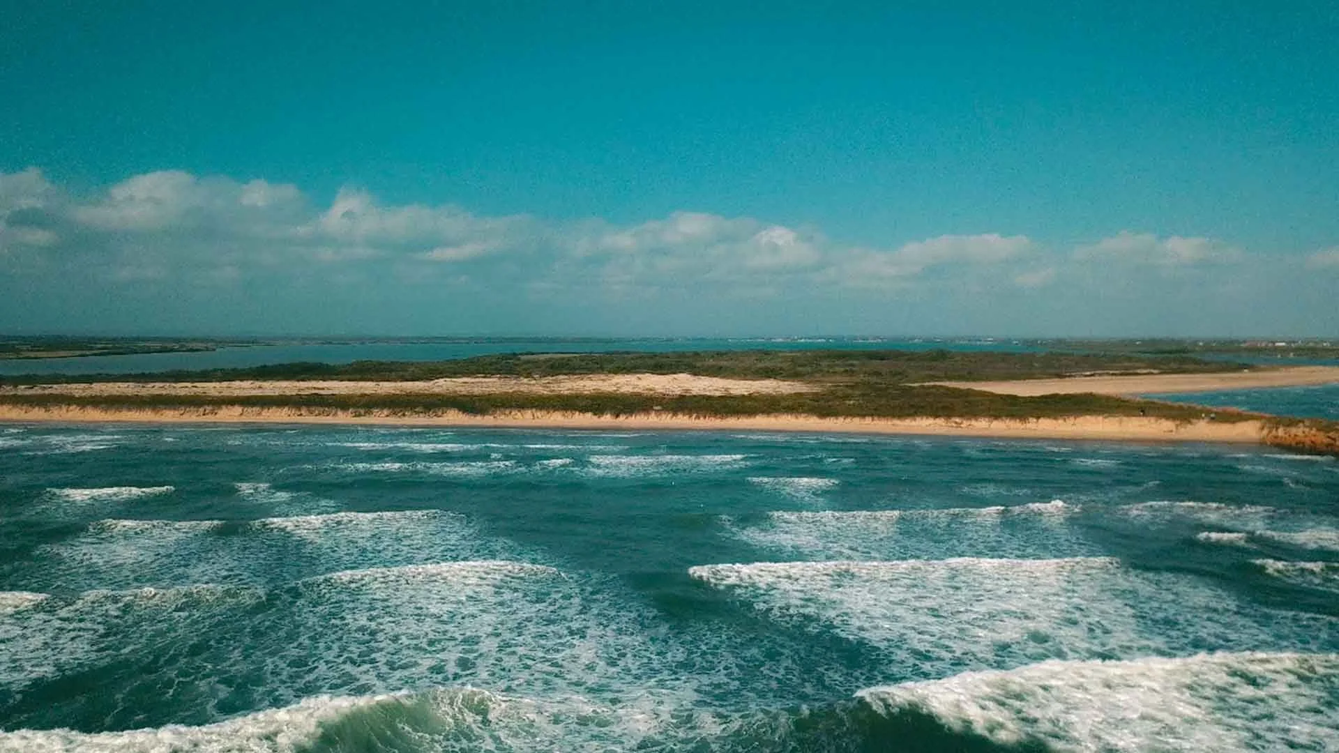 Aerial view of a coastal landscape with ocean waves in the foreground, sandy dunes in the middle, and a landmass with shrubs and trees in the background under a partly cloudy sky.