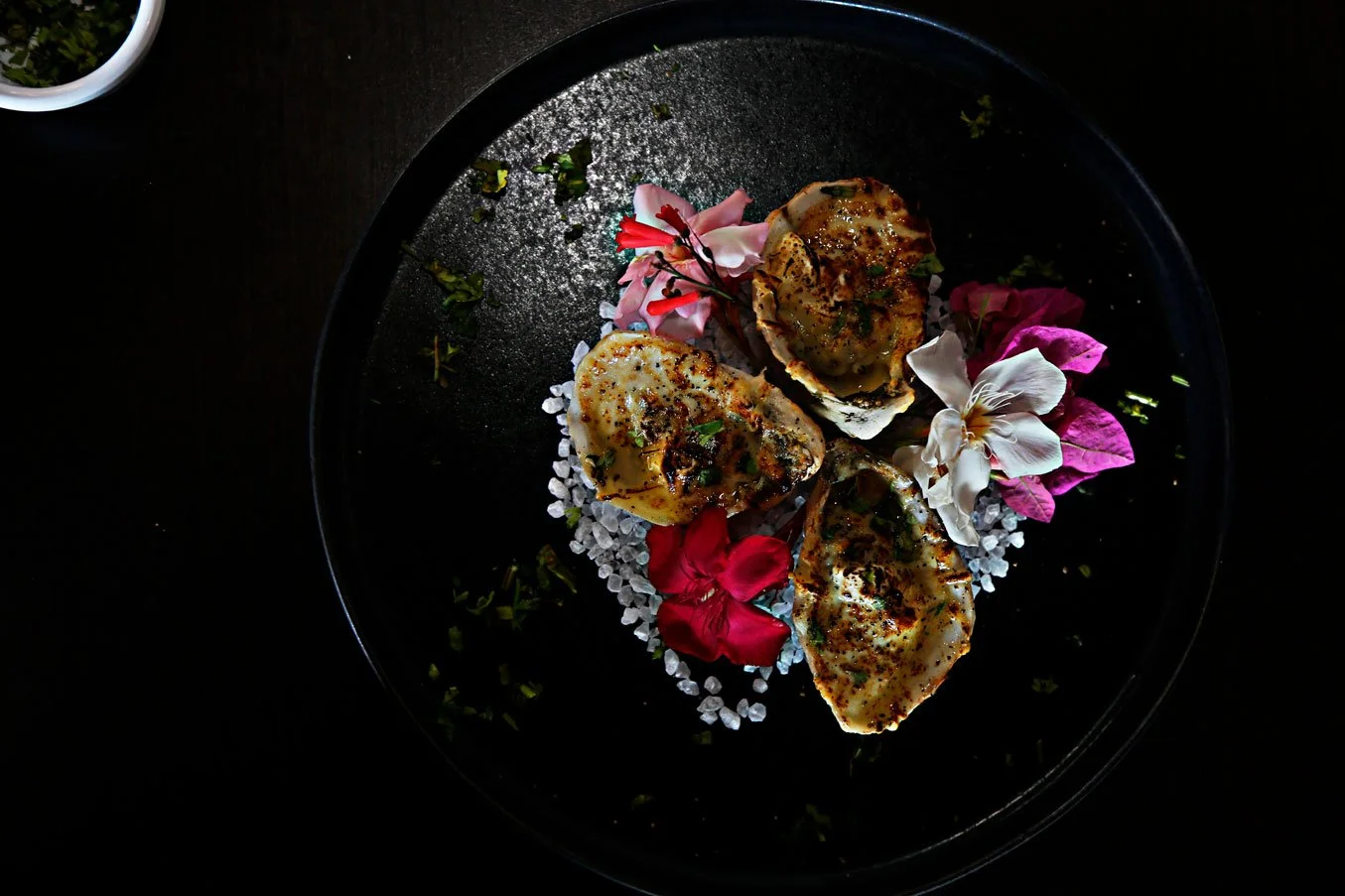 Three baked oysters garnished with herbs, arranged on a bed of coarse salt crystals, surrounded by pink, white, and purple flowers, on a black plate.