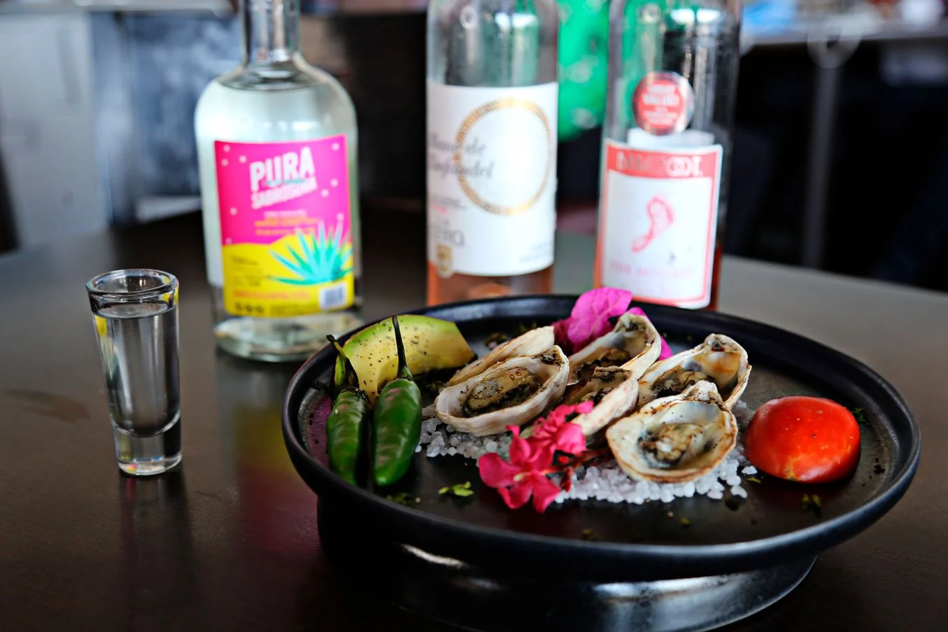 A plate of raw oysters on ice with a slice of melon, two green chili peppers, a tomato, and pink flowers. Behind the plate, there are three bottles of alcohol and a shot glass with clear liquid on a dark table.