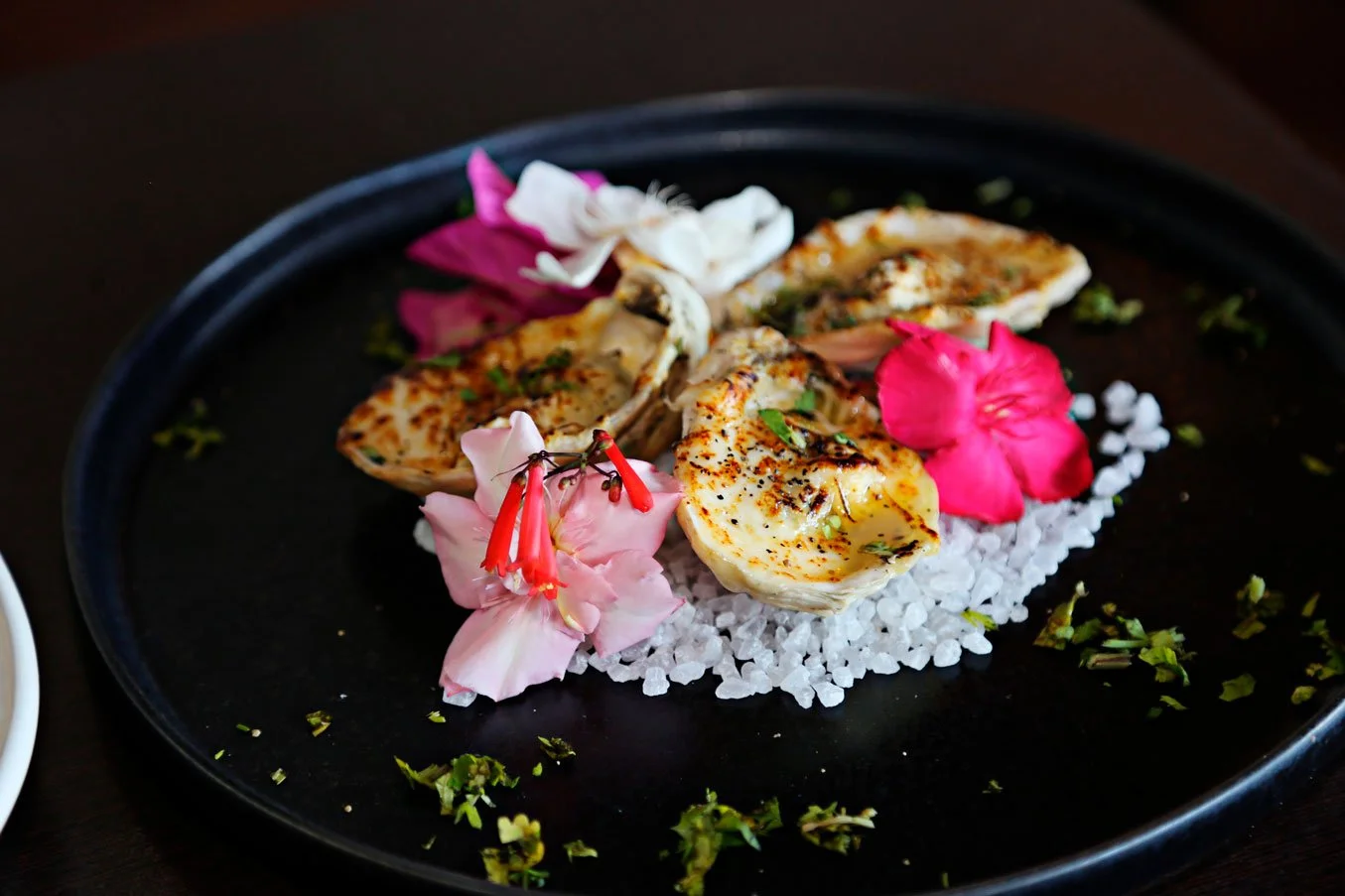 Grilled oysters garnished with herbs, served on a black plate with pink and white flowers and sea salt.