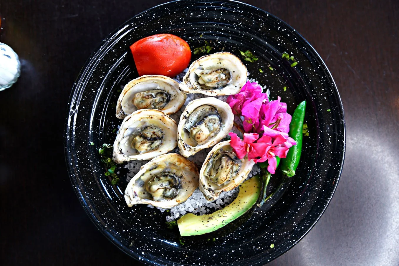 A black speckled plate with six oysters on the half shell, a cherry tomato, a slice of avocado, a green chili, and pink edible flowers, all arranged on a bed of salt.
