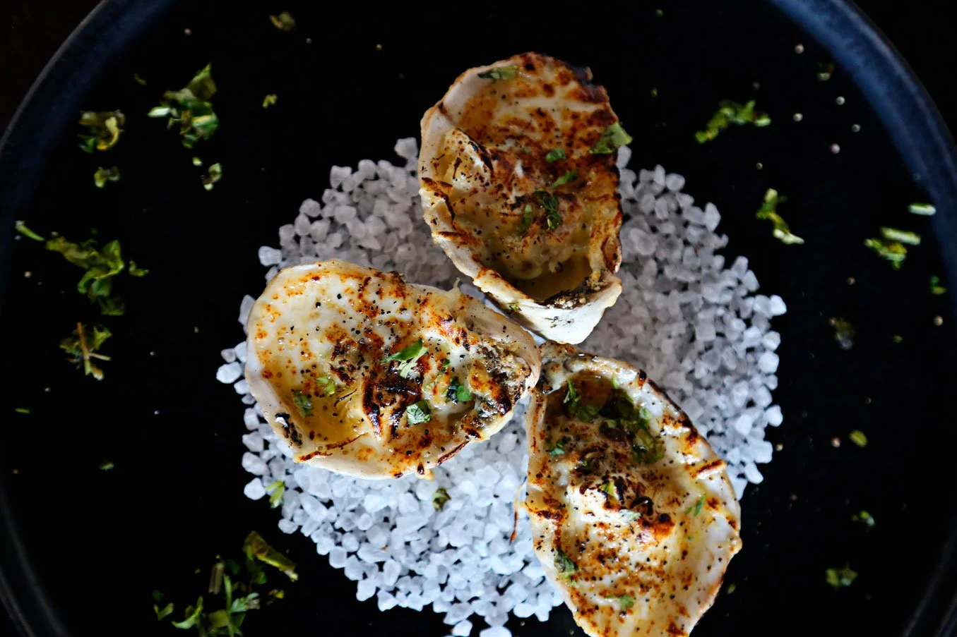 Three grilled oysters garnished with herbs, served on flaky salt with scattered herbs on black plate.