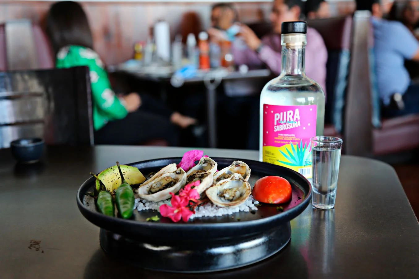 A black plate with oysters garnished with lime, cucumber, and pink flowers on a salt bed, placed on a table. In the background, there is a bottle of flavored liquor labeled "Pura Sabrosa" and a shot glass filled with clear liquid, with blurred restau