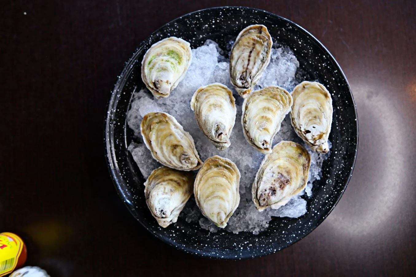 A black speckled bowl filled with ice and fresh oysters on a dark wooden surface.
