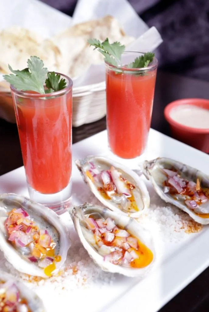 Seafood oysters on a bed of salt with chopped onions, alongside two glasses of red drink garnished with cilantro, and a basket of bread in the background.