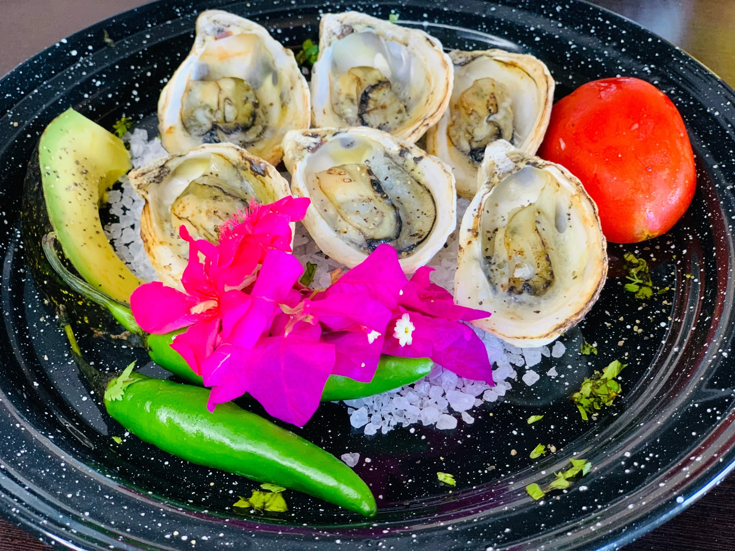 A plate of fresh oysters on the half shell garnished with pink flowers, a slice of avocado, a slice of tomato, and green chili, all served on a bed of salt with a black speckled tray.