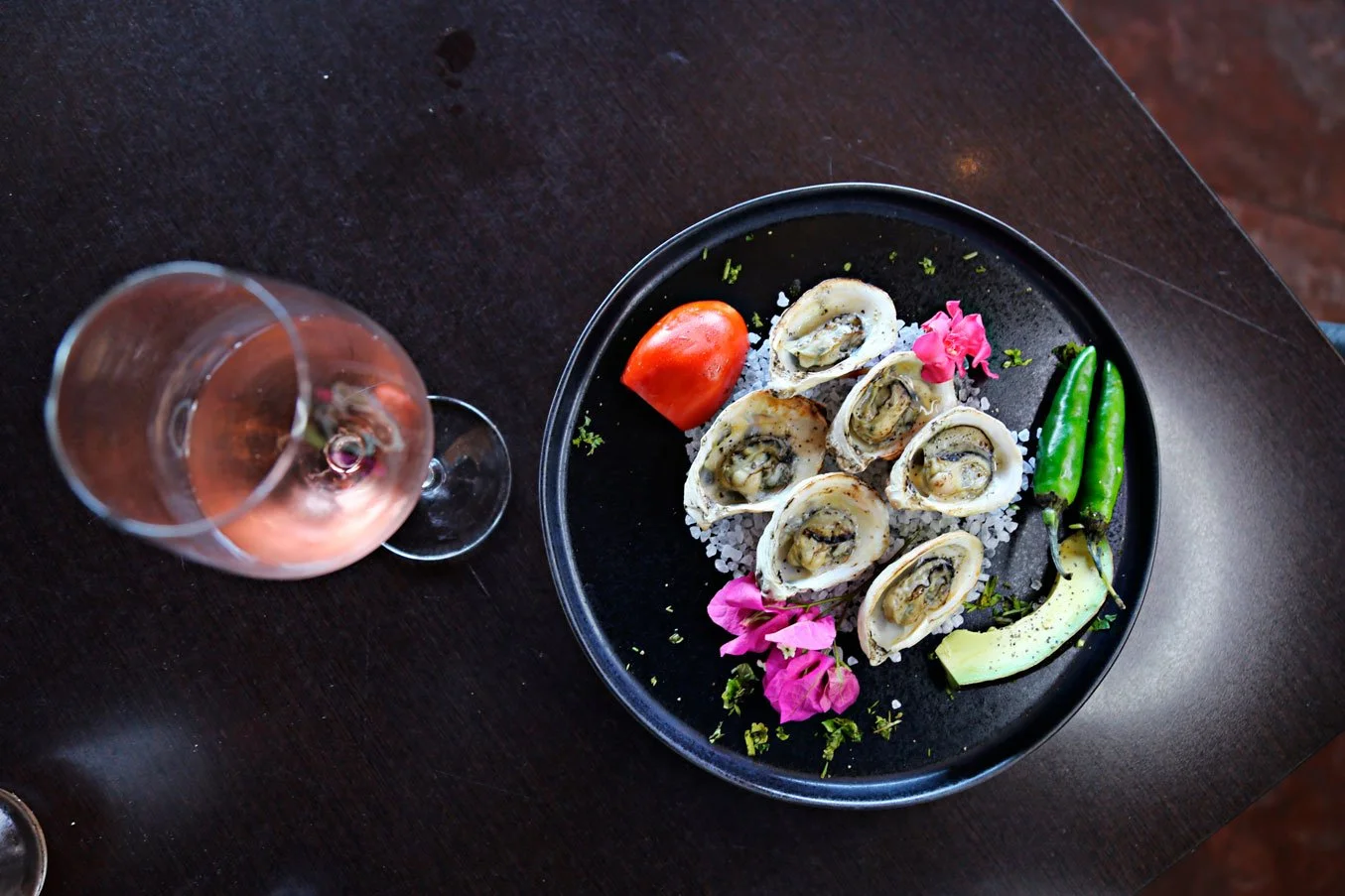 A black plate with oysters on a bed of salt, garnished with pink flowers, cherry tomato, sliced avocado, and two green chili peppers. A glass of rosé wine is placed nearby on a dark wooden table.
