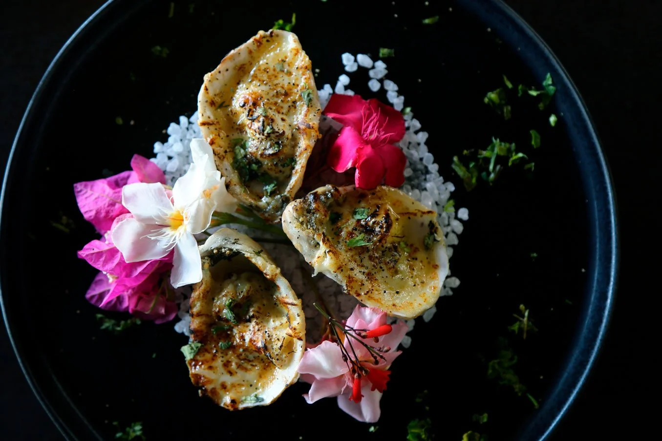 Three grilled oysters on a black plate garnished with edible flowers and herbs