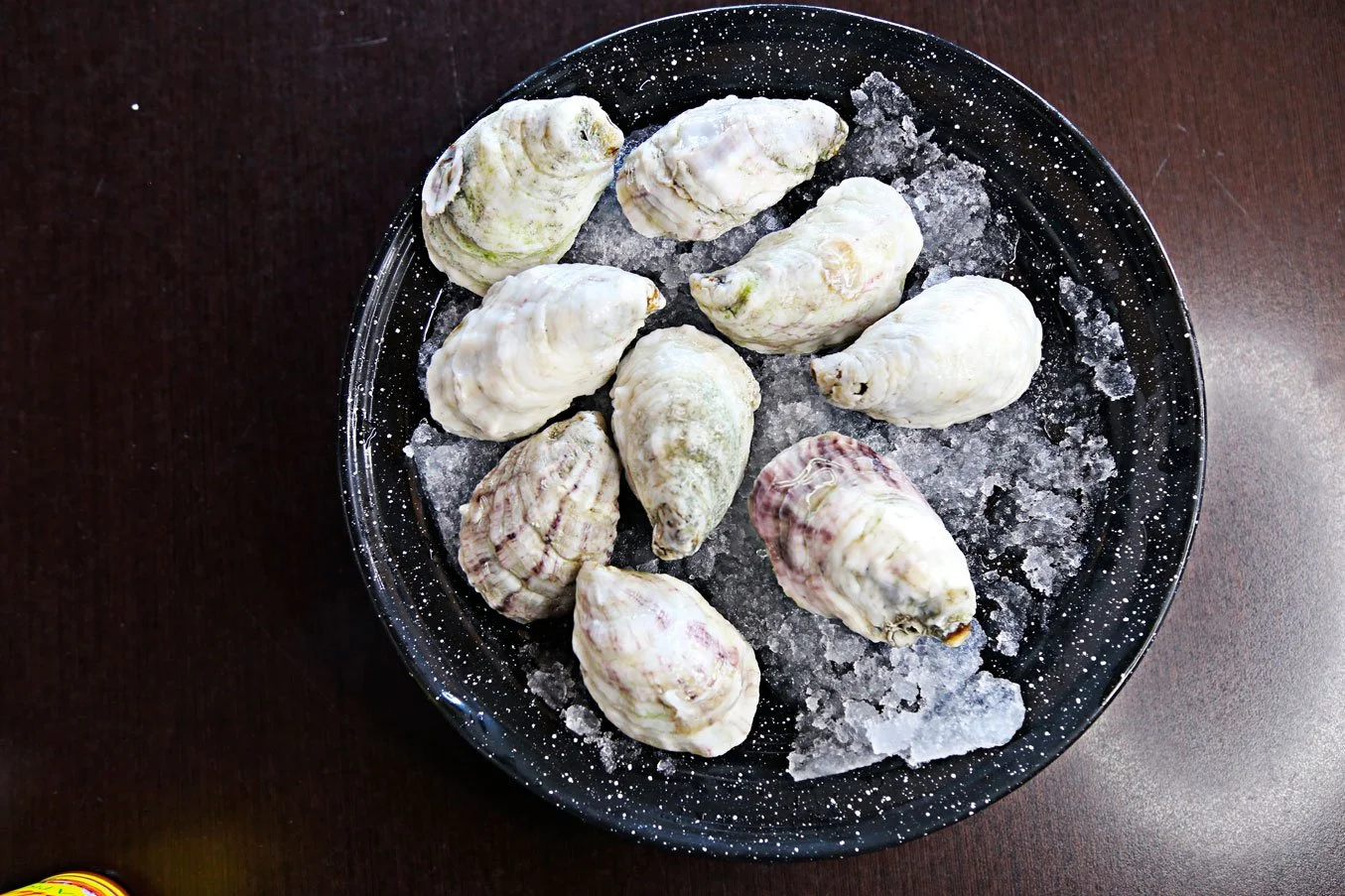 A black speckled bowl with ice and several raw, shelled oysters on top, on a dark brown surface.