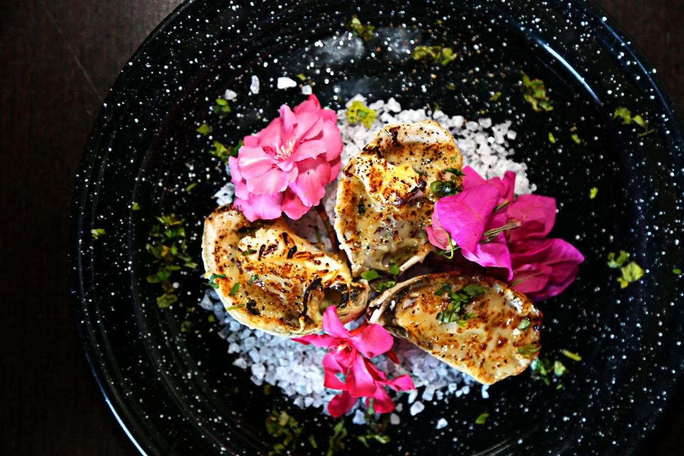 Grilled oysters topped with herbs and spices, garnished with pink flowers, on a black speckled plate with white salt flakes.