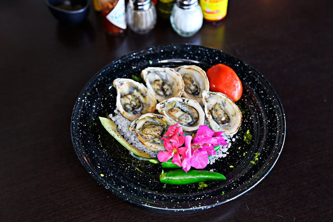 A black speckled plate with six oysters on the half shell, a small tomato, pink flowers, a green chili, and cucumber slices, set on a dark wooden table.