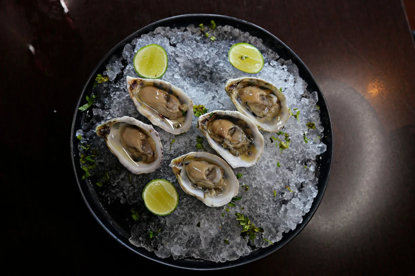 A black plate with six fresh oysters on ice, garnished with chopped herbs and three lime wedges.