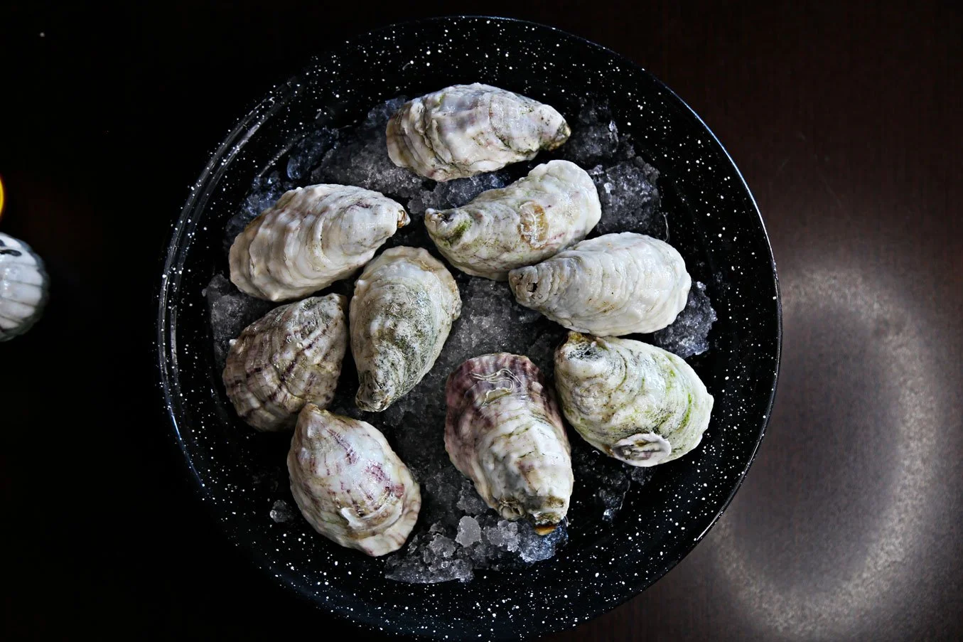 A black speckled bowl containing raw oysters on a dark surface.