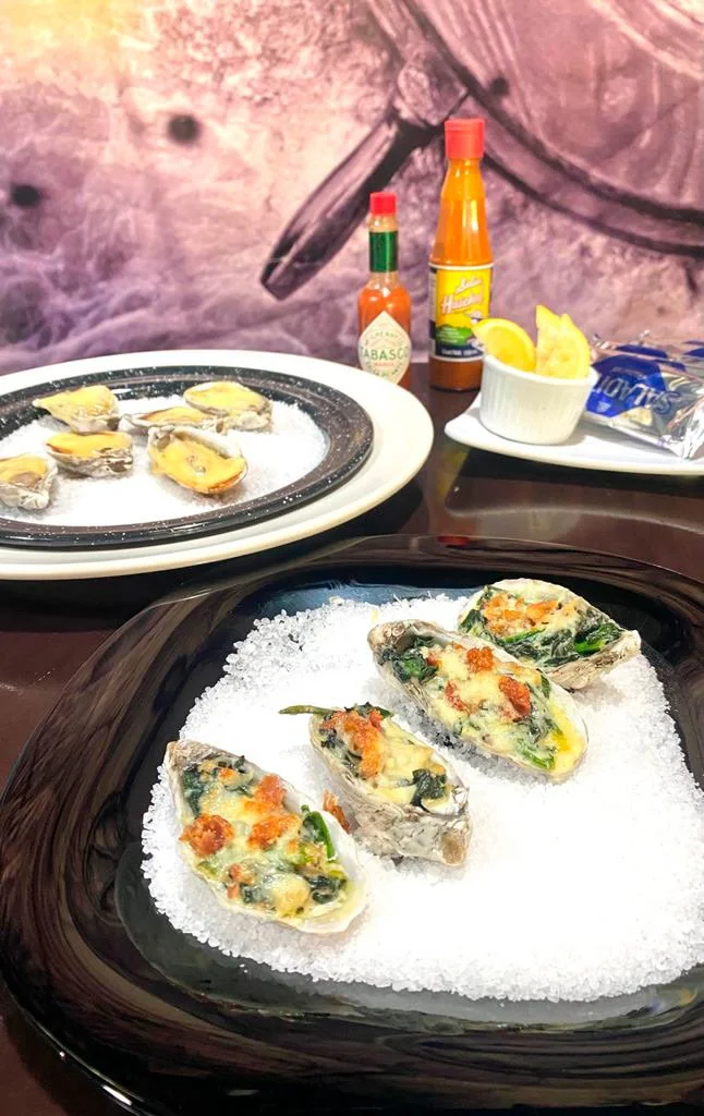 A plate of raw oysters topped with herbs and cheese, and another plate with cooked oysters, alongside hot sauce, lemon slices, and a snack package on a dark table with a pinkish background.