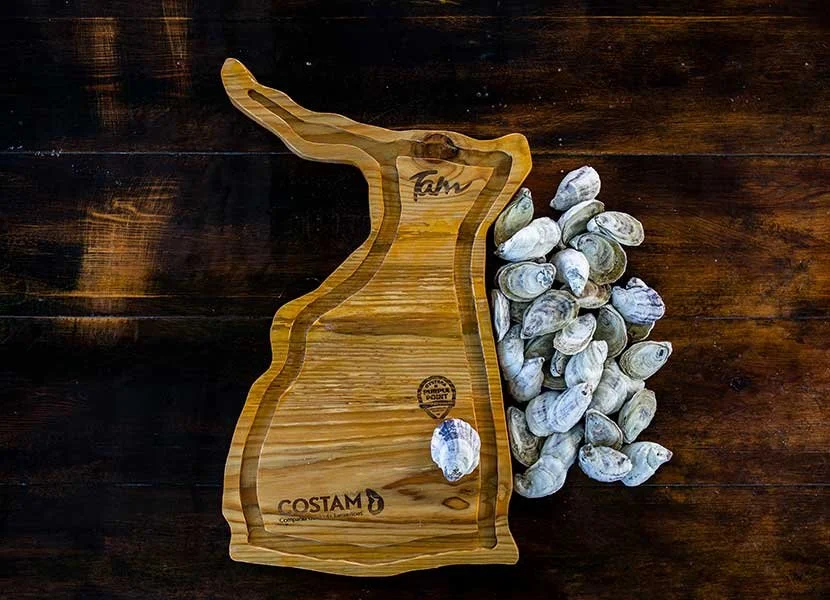 Wooden tray shaped like the state of Georgia filled with oysters, with more oysters on the side, on a dark wooden surface.