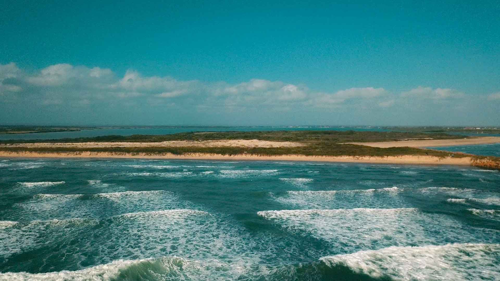Aerial view of a coastal landscape with sandy beaches, green vegetation, and blue ocean waves under a partly cloudy sky.