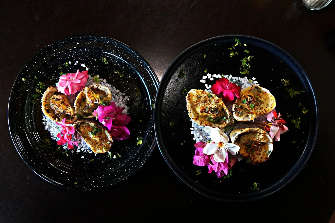Two black plates with oysters garnished with pink flowers, salt, herbs, and spices on a dark surface.