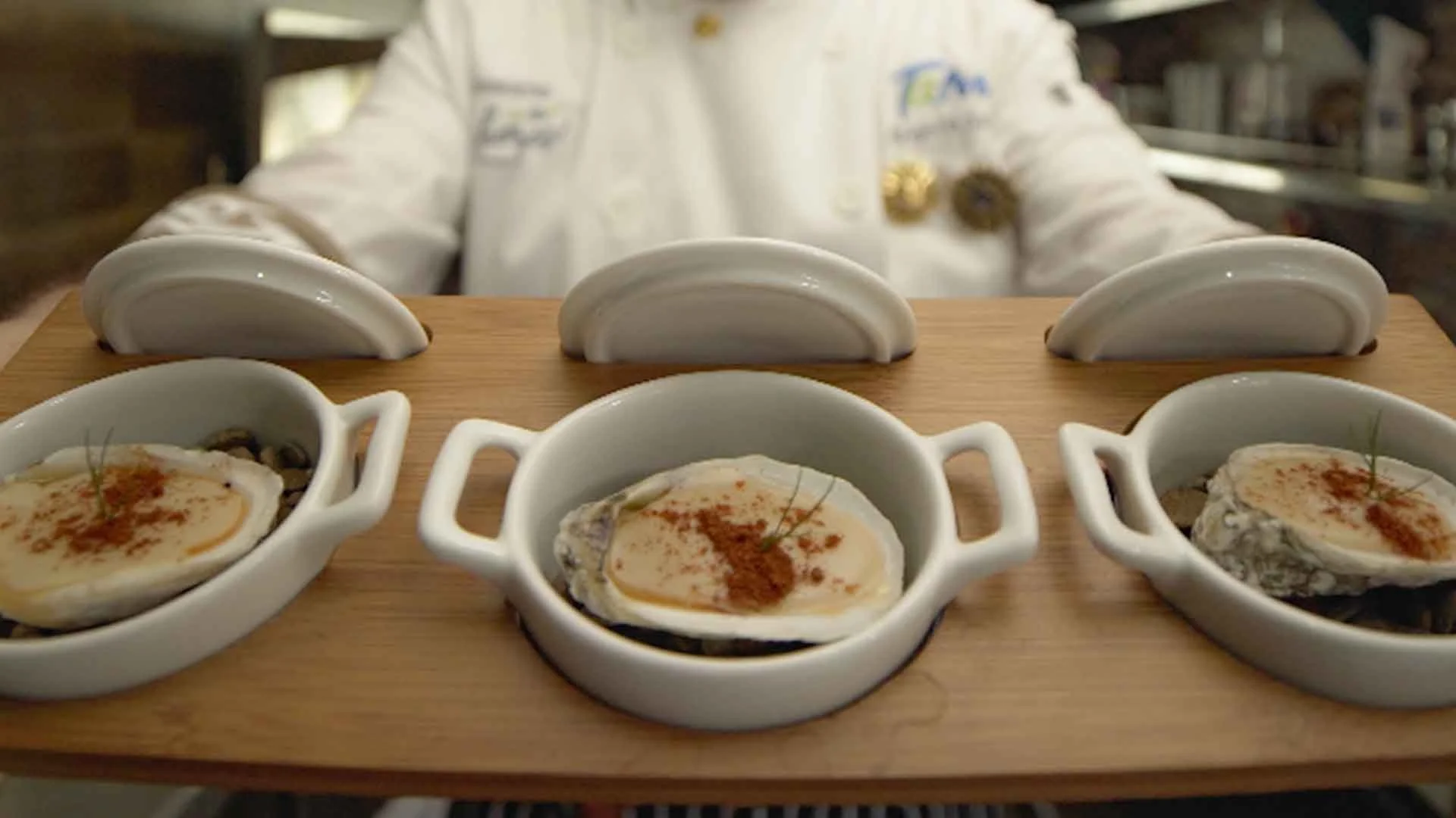 Dish with three cooked oysters topped with seasoning and garnished with herbs, served in small white ramekins on a wooden platter. Behind the platter, a person in a chef's uniform is preparing the dish.
