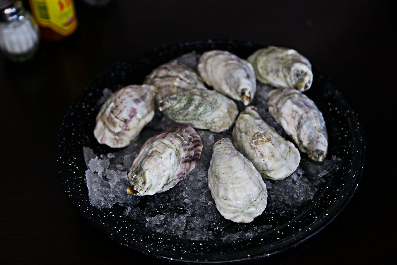 A black platter filled with fresh oysters on ice, with salt grains sprinkled around, on a dark table, with some condiments visible in the background.