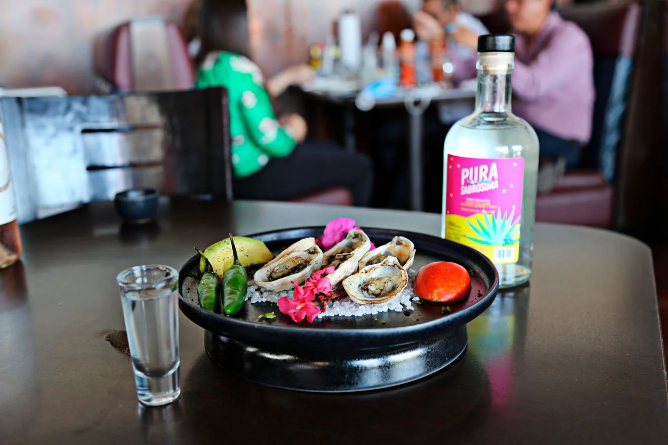 A black circular plate with oysters on ice, garnished with pink flowers, lime, green chili peppers, a tomato, a small bottle of tequila with a colorful label, and a glass of clear liquid on a restaurant table. Blurred background of people dining.