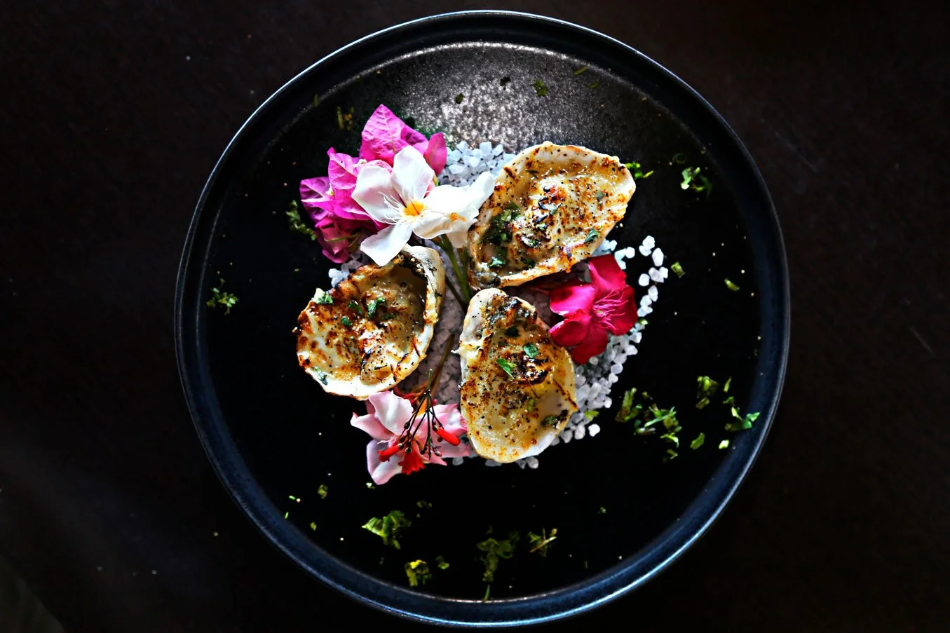 Scallops garnished with herbs and spices, served on a black plate, topped with edible flowers and sea salt