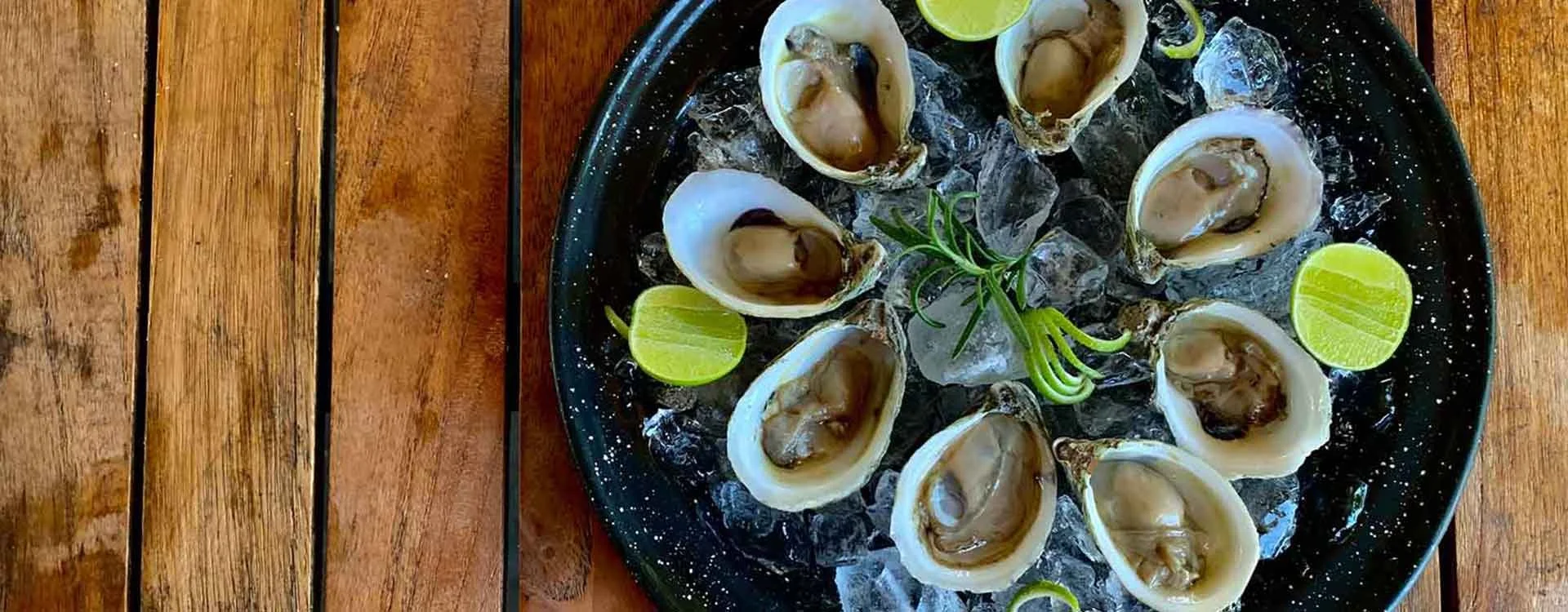 A black plate filled with ice and fresh oysters garnished with lime wedges and a sprig of rosemary on a wooden table.