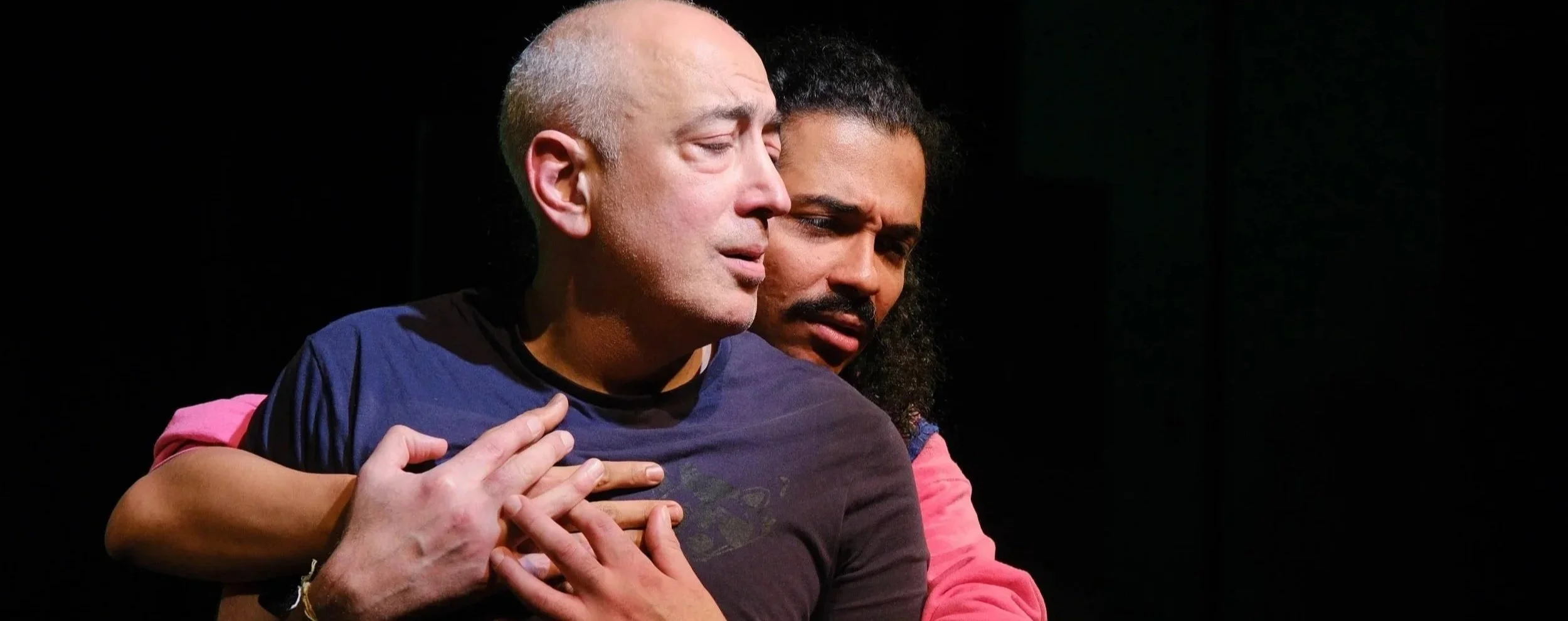 Two men sharing an emotional moment, one resting his hand on the other's chest, both appearing somber with eyes closed or looking downward, against a dark background.