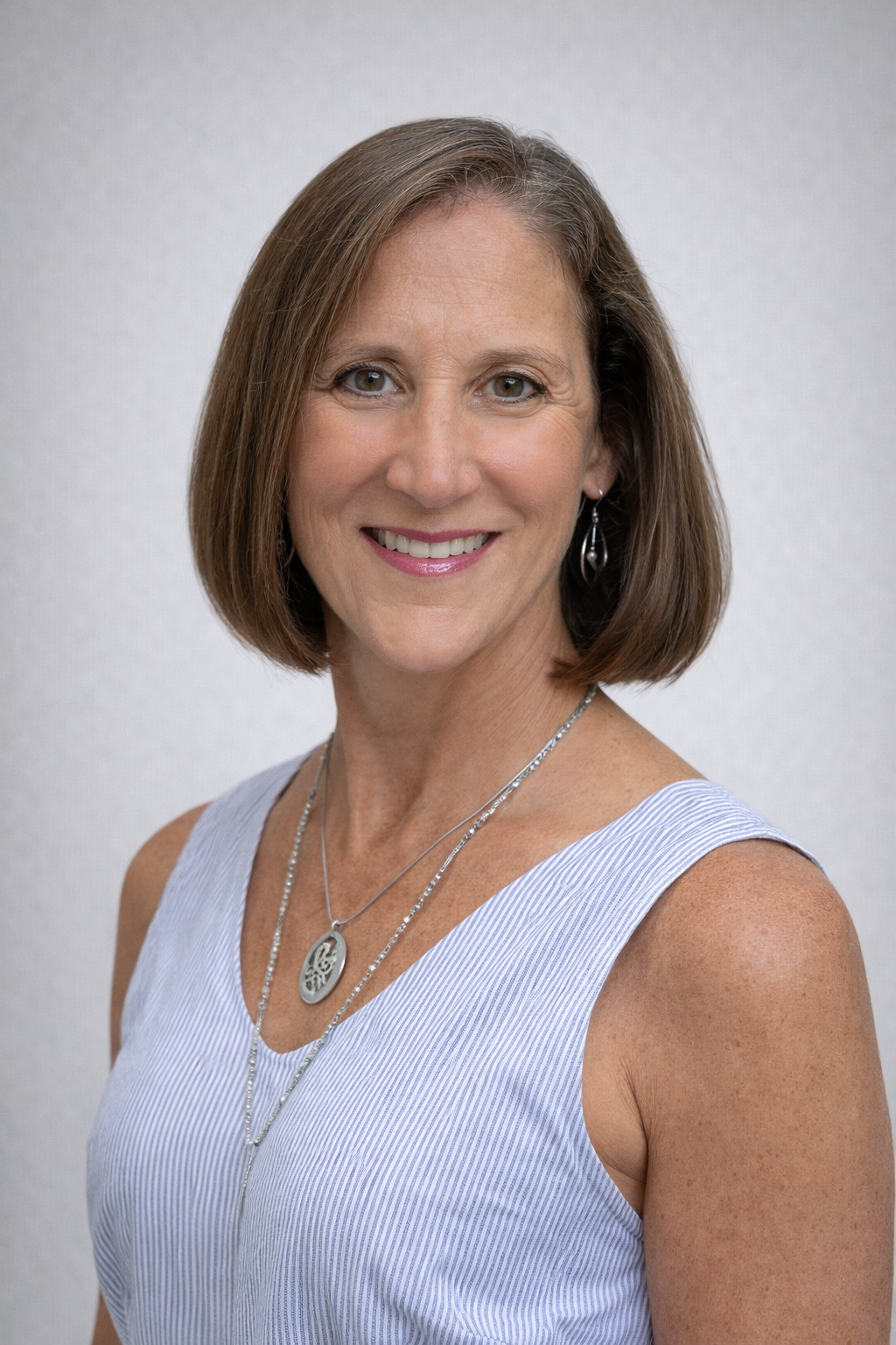 A smiling woman with shoulder-length brown hair, wearing a light blue and white striped sleeveless top, silver earrings, and layered silver necklaces, standing against a plain light gray background.