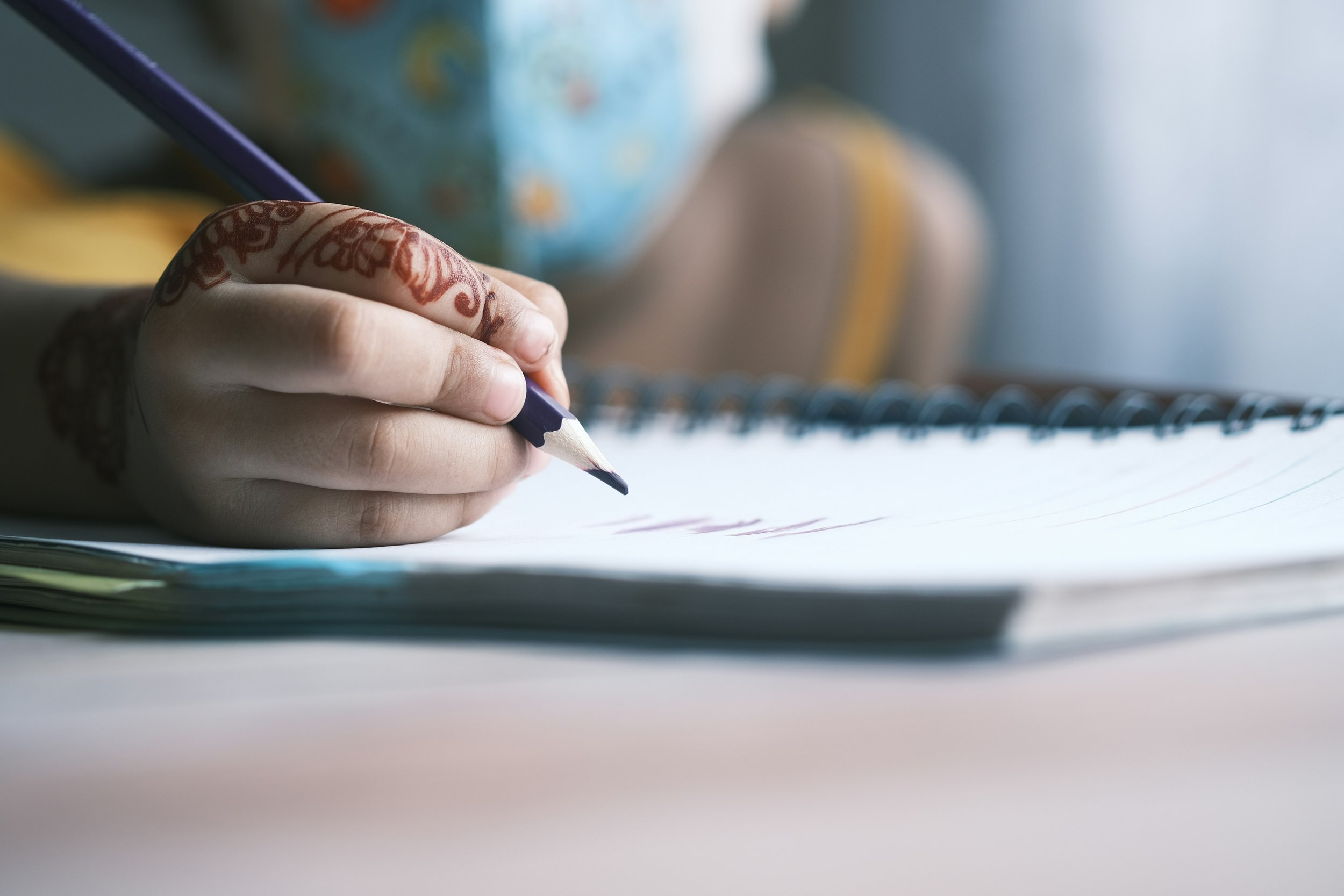 Close-up of a hand holding a dark purple colored colored pencil, writing on a spiral-bound notebook with white pages. The person has intricate henna designs on their fingers and thumb. The background is blurred.
