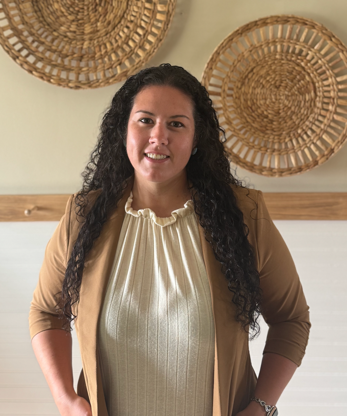 A woman with long curly black hair smiling, wearing a beige blazer over a cream-colored pleated blouse, standing in front of woven basket wall art.