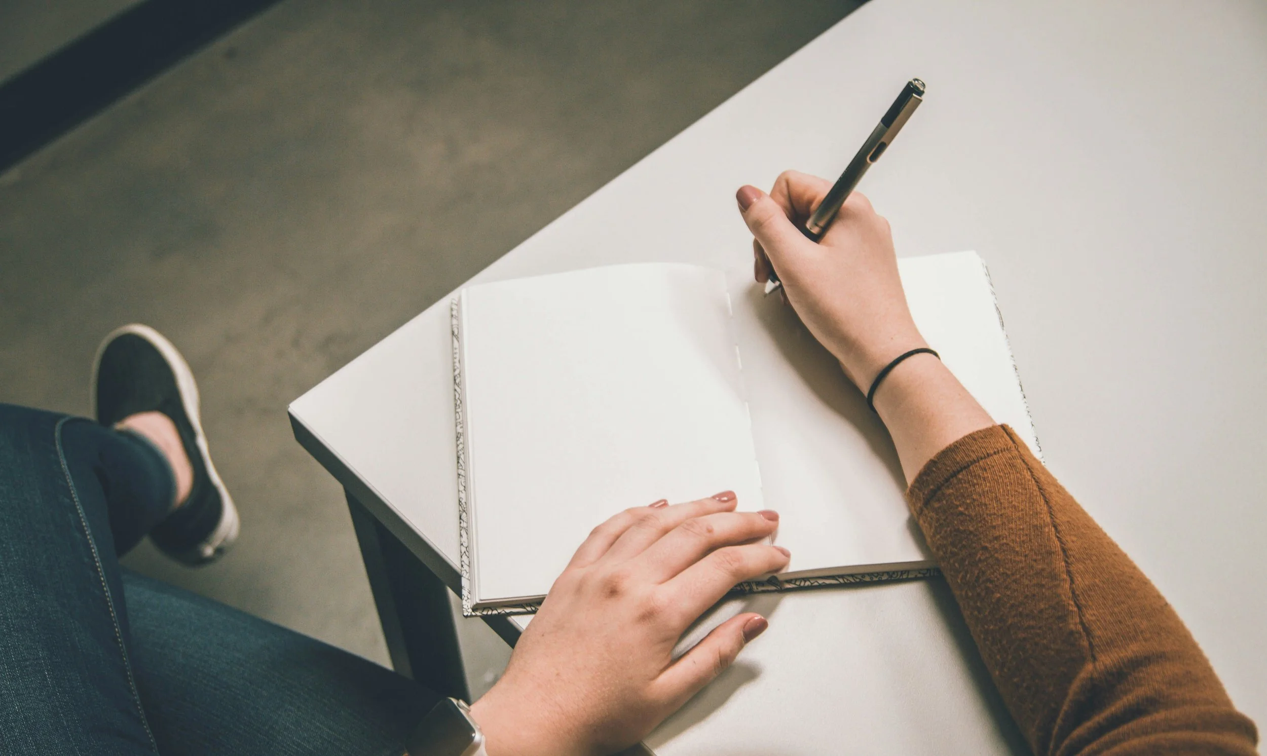 A person writing in a notebook with a black pen on a white table, wearing a brown sweater, with another person's hand resting on the notebook and a wristwatch visible.
