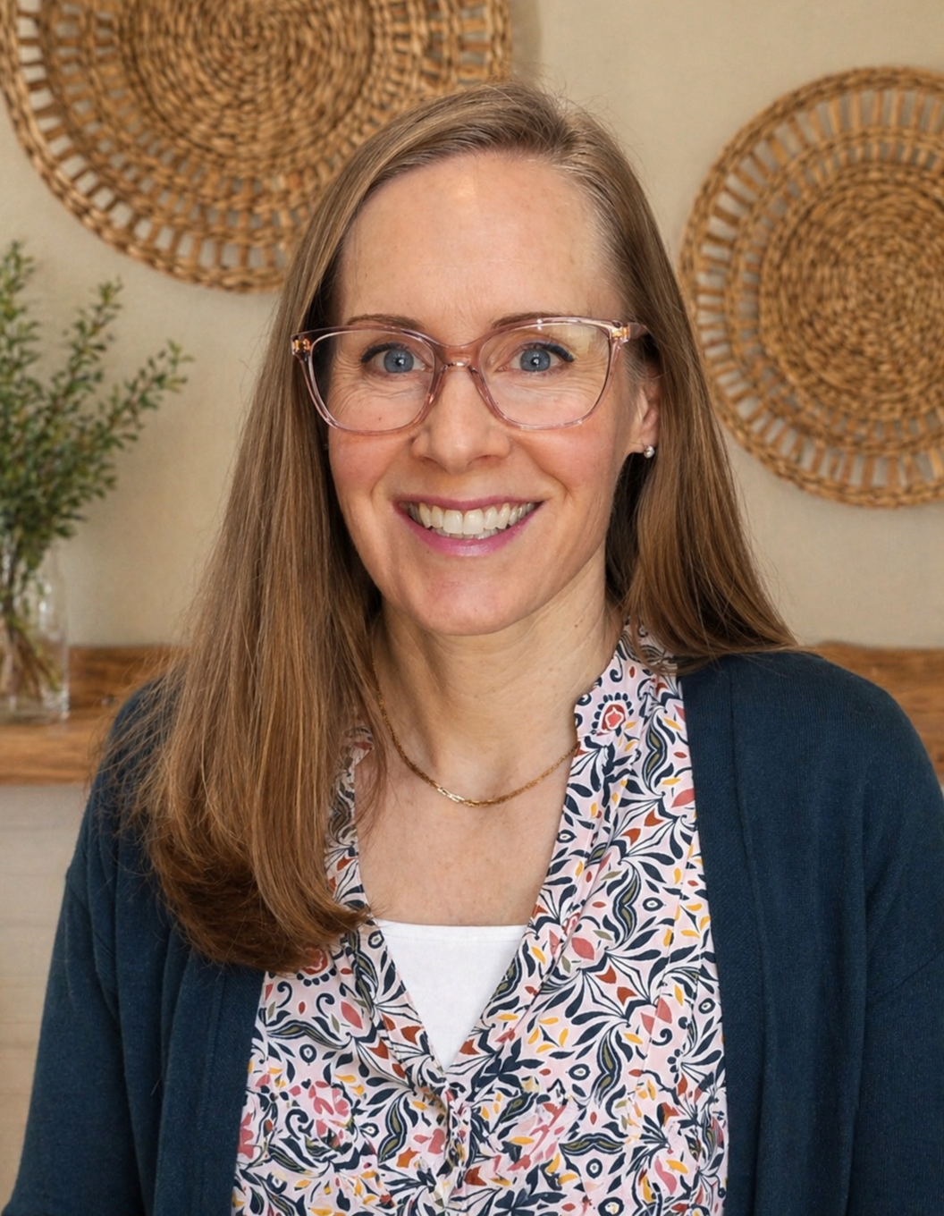 A woman with long red hair, wearing pink glasses, a patterned blouse, and a navy cardigan, smiling at the camera in a cozy room with woven wall hangings and a plant.