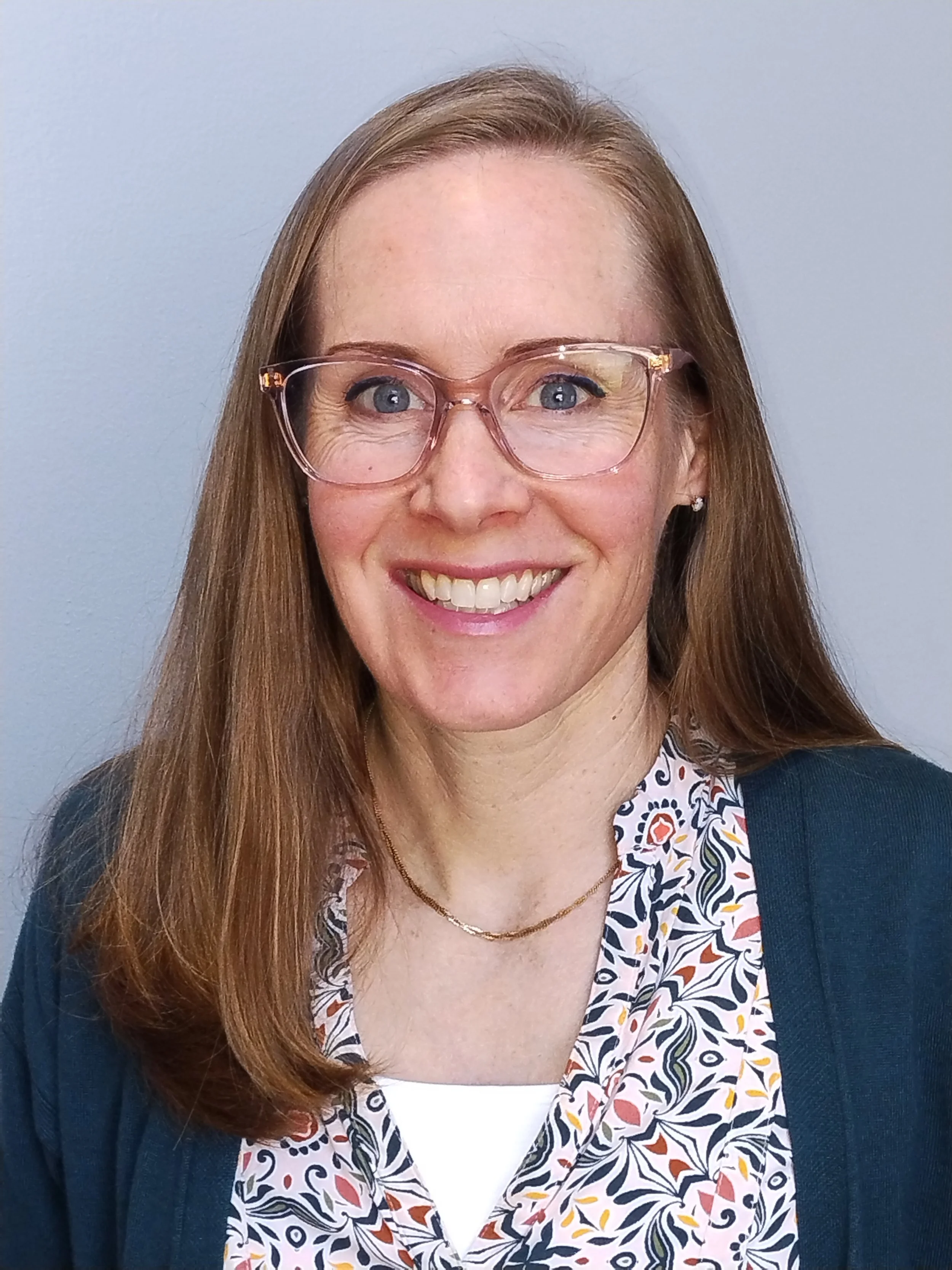 Close-up of a woman with long red hair, wearing large clear eyeglasses, floral blouse, navy blazer, small earrings, and a gold necklace, smiling against a gray background.
