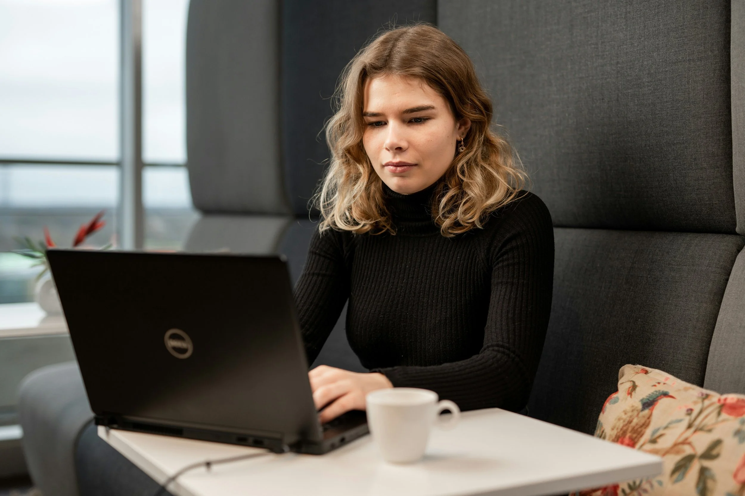 A young woman with wavy blonde hair wearing a black turtleneck sitting at a table in a modern office, looking at her laptop, with a white mug and a floral-patterned bag on the table.