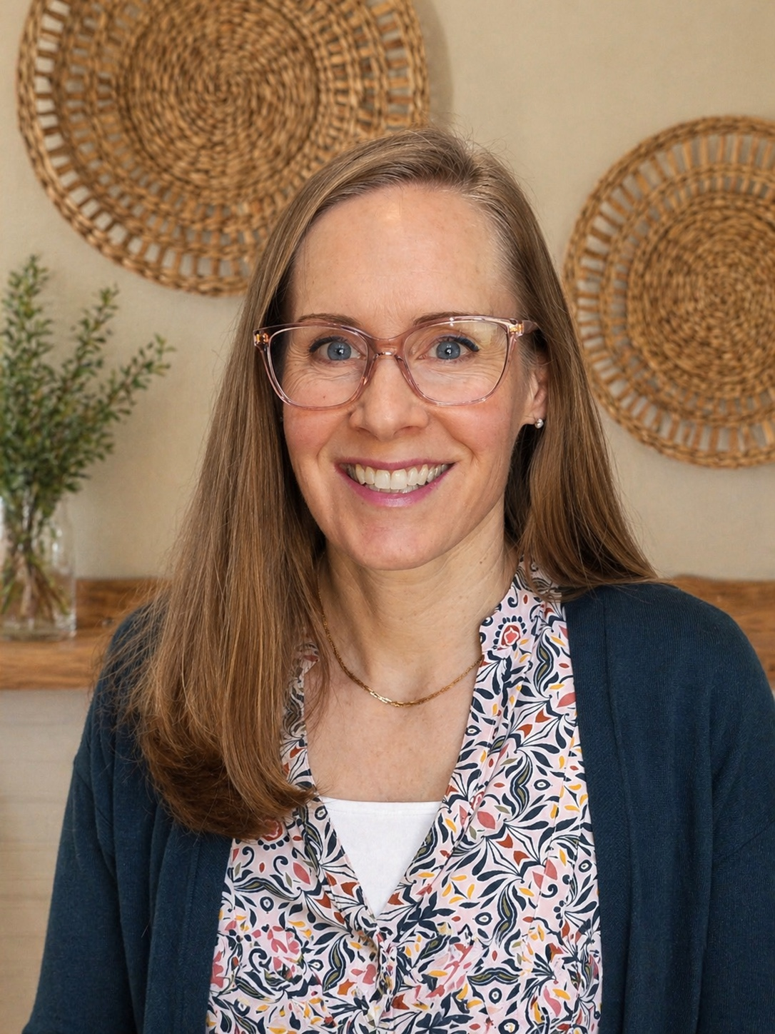 A woman with long brown hair and glasses smiling at the camera, wearing a patterned blouse and a dark cardigan, with decorative woven wall hangings and a potted plant in the background.