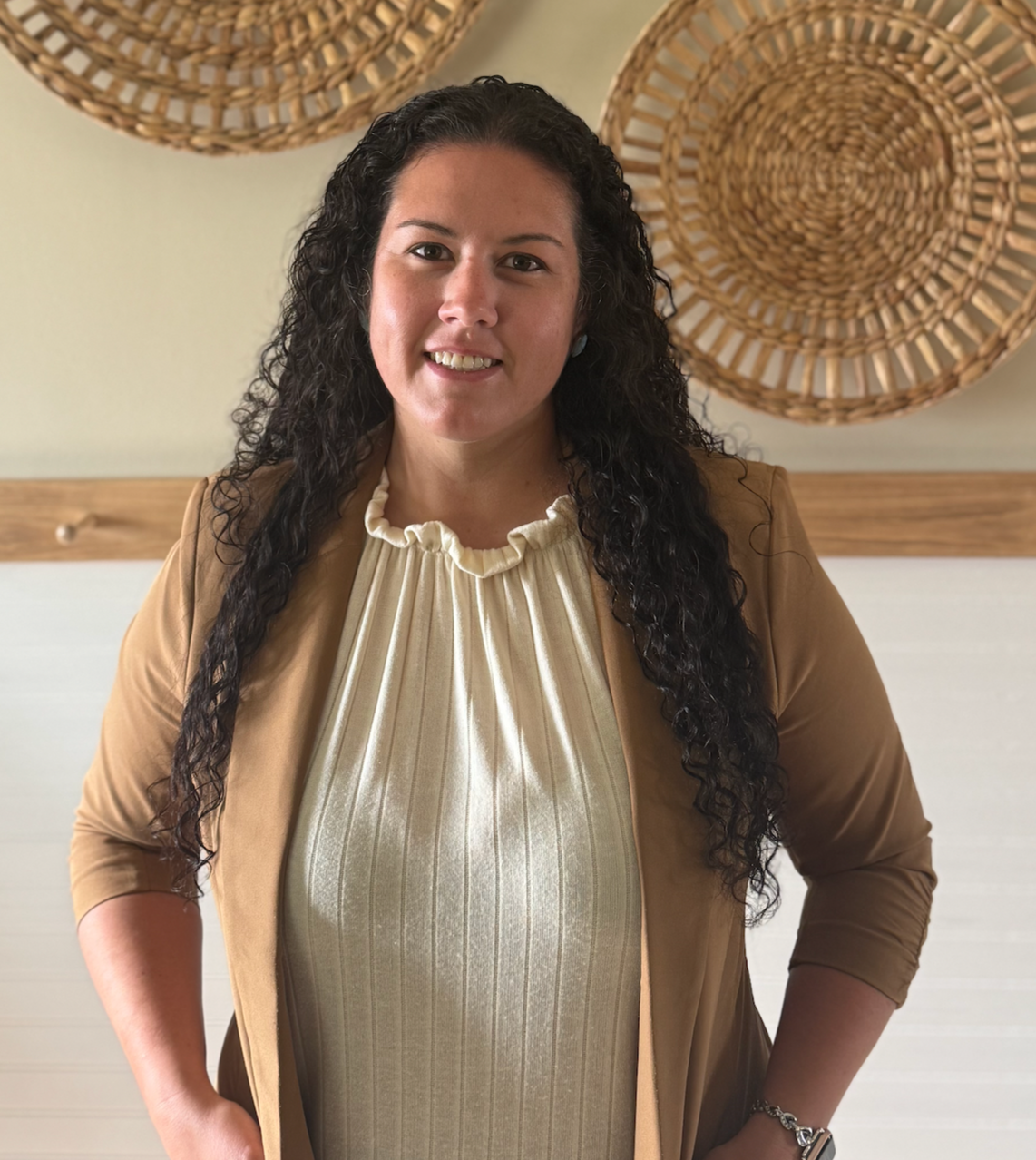 A woman with long curly dark hair smiling at the camera, wearing a beige blazer and a cream ruffled blouse, standing in front of a wall decorated with woven round wall hangings.