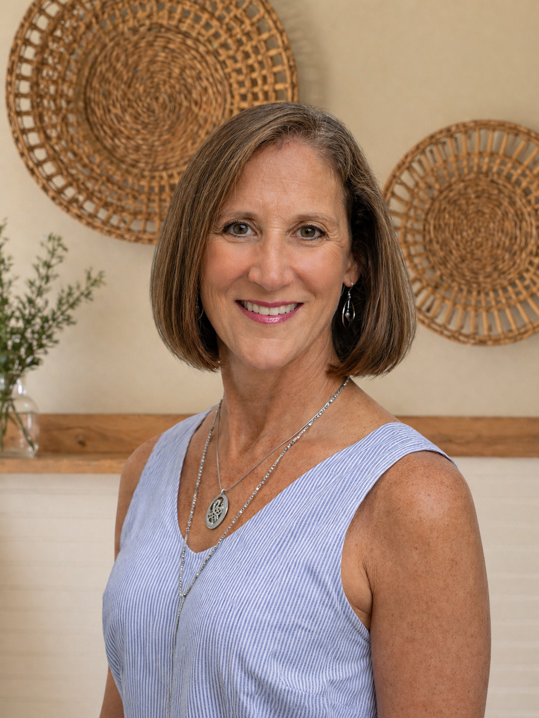 A woman with shoulder-length brown hair smiling, wearing a sleeveless blue and white striped top, silver jewelry, and earrings, standing in front of a beige wall with woven decorations and green plant.