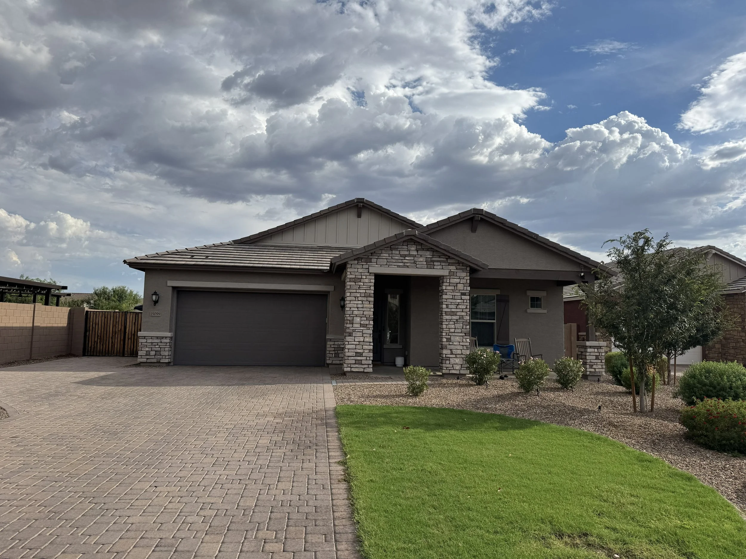 A residential house with stone and stucco exterior, a dark tiled roof, a garage door, and a driveway. The front yard has a paved walkway and green grass, with a fence on the left and a tree on the right. The sky has scattered clouds with patches of blue.