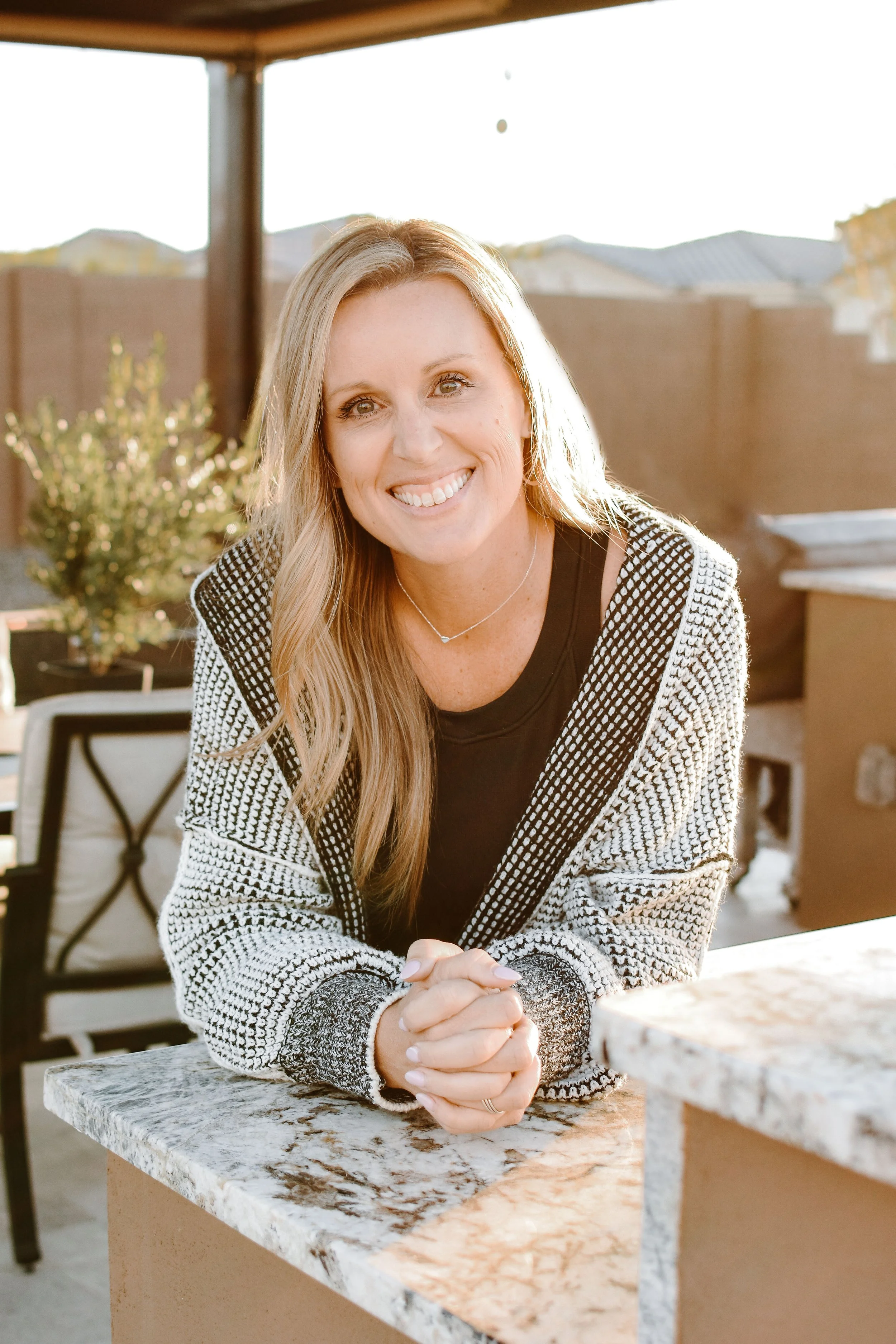 A woman with long blonde hair smiling and sitting at an outdoor marble table on a patio, with a wooden fence and rooftops in the background during the daytime.