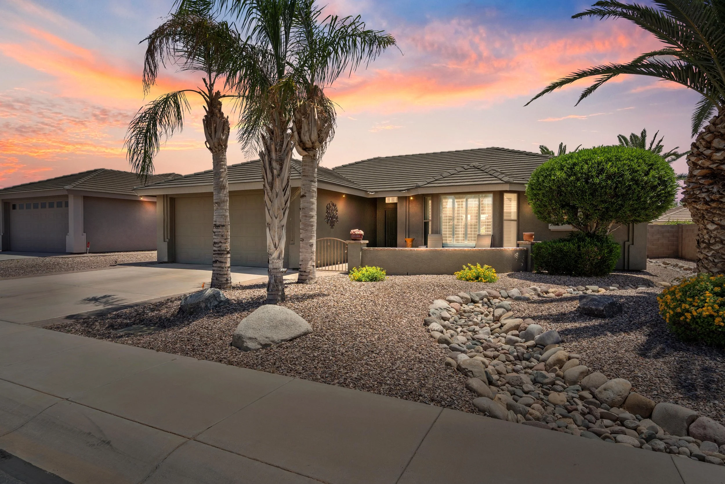 A suburban house at sunset with a tiled roof, palm trees, and desert landscaping including rocks, yellow flowers, and a small tree.