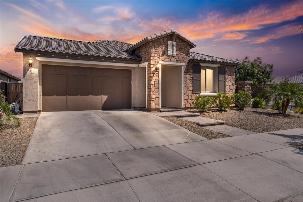 Front view of a house with a stone facade, a two-car garage, some small trees and bushes, a pathway, and a sunset sky in the background.