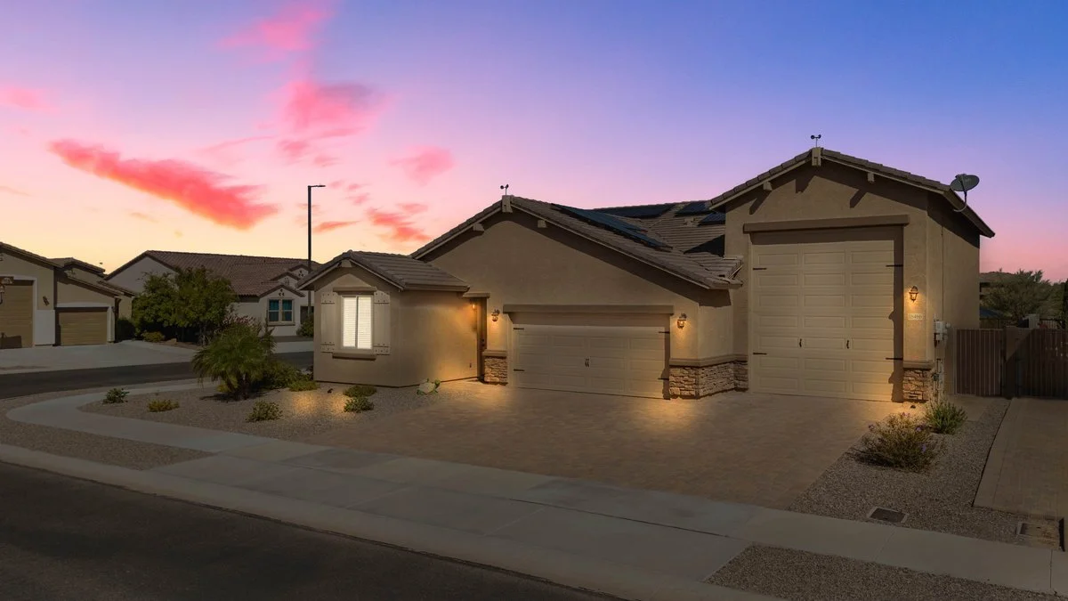 A single-story house with a two-car garage, front yard landscaping with small bushes and trees, sunset sky with pink and purple clouds.
